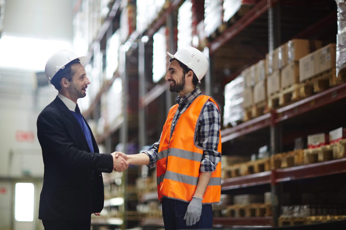 Happy foreman in helmet greeting one of warehouse workers